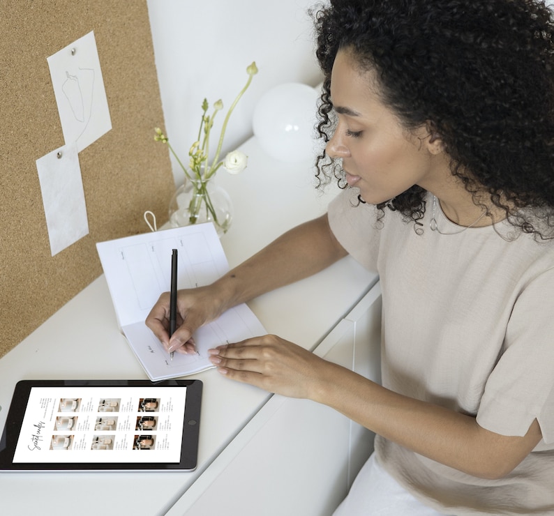 Puede incluir: Una persona escribe en una agenda en un escritorio blanco con un bol&iacute;grafo negro. Una tableta que muestra im&aacute;genes est&aacute; en el escritorio. Un corcho con bocetos y un jarr&oacute;n con flores est&aacute;n en el fondo. La persona lleva una camiseta beige.