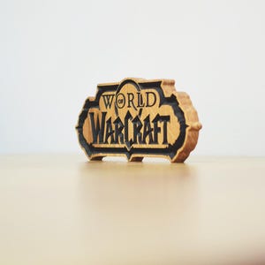 May include: A wooden sign with the words "World of Warcraft" carved into it. The sign is in a stylized font with a black outline and a light brown wood grain background. The sign is on a light brown surface.