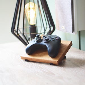 May include: A black games controller sits on a wooden stand. The stand is made of light-coloured wood and has a slanted design. A black geometric lamp and a speaker are in the background.