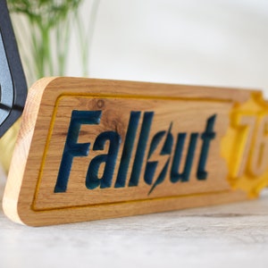 Op de afbeelding: Een houten bord met de woorden "Fallout 76" in blauwe en gele verf. Het bord is gemaakt van lichtbruin hout en heeft een gele rand.