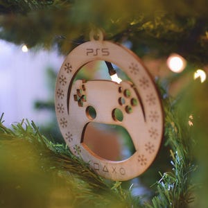 Op de afbeelding: Een houten kerstversiering in de vorm van een PS5-controller. De lichtbruine versiering heeft de letters "PS5" bovenaan, sneeuwvlokpatronen langs de rand en een gedetailleerd controllerontwerp met knoppen en symbolen.