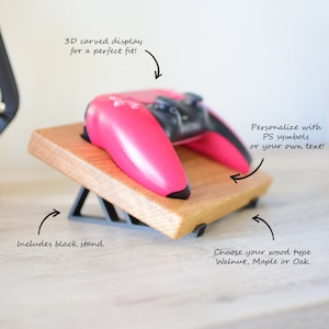 Peut inclure: Une manette de jeu vidéo rose est posée sur un support en bois avec une base en métal noir. Le support est incliné vers le haut. Le texte "3D carved display for a perfect fit!" est au-dessus de la manette. Le texte "Personalize with PS symbols or your own text!" est à droite de la manette. Le texte "Includes black stand" est sous le support. Le texte "Choose your wood type Walnut, Maple or Oak" est sous le support.