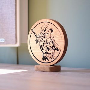 Può includere: Un pezzo decorativo in legno, circolare, con un'immagine incisa in nero di un personaggio della serie di videogiochi The Legend of Zelda. Il personaggio è raffigurato con una spada e uno scudo. Il pezzo è montato su una piccola base di legno.