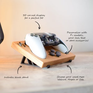 Peut inclure: Une manette de jeu vidéo blanche et noire est posée sur un support en bois avec une base en métal noir. Le support est incliné vers le haut et présente un design sculpté en 3D. Le texte "Personnaliser avec des symboles Ps, votre propre texte ou des accessoires de magasin !" est écrit en blanc sur fond noir. Le texte "Comprend un support noir" est écrit en blanc sur fond noir. Le texte "Choisissez votre type de bois Noyer, Érable ou Chêne" est écrit en blanc sur fond noir.
