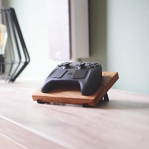 Pu&ograve; includere: Un controller per videogiochi nero appoggiato su un supporto in legno inclinato. Il supporto presenta una finitura con venature del legno naturali ed &egrave; sostenuto da piccole staffe metalliche nere alla base per un'esposizione angolata.