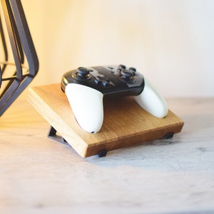 Op de afbeelding: Een houten standaard met een zwart-witte videogamecontroller erop. De standaard heeft een driehoekige basis en is gemaakt van lichtbruin hout.