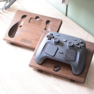 Pu&ograve; includere: Un supporto per controller in legno marrone scuro con un intaglio per un controller di gioco e il logo Steam inciso sulla parte anteriore. Una seconda base in legno abbinata con il testo VALVE inciso &egrave; posizionata dietro il supporto principale.