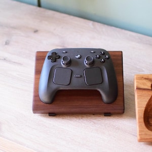 Pu&ograve; includere: Un controller per videogiochi nero appoggiato su un supporto rettangolare in legno marrone scuro. Il controller presenta una croce direzionale, due levette analogiche, quattro pulsanti azione etichettati X, Y, A e B e un tasto centrale Steam.