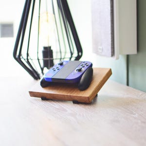 Op de afbeelding: Een blauwzwarte gamecontroller ligt op een houten standaard. De standaard is gemaakt van lichtbruin hout en heeft zwarte steunen. Een zwarte geometrische lamp en een witte luidspreker staan op de achtergrond.