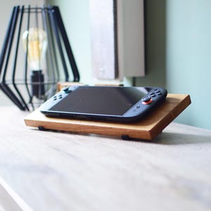 Op de afbeelding: Een zwarte handheld gameconsole ligt op een houten standaard met twee niveaus. De standaard is gemaakt van lichtbruin hout en heeft een rechthoekige vorm. De console heeft zwarte knoppen en een zwart scherm. Een zwarte geometrische lamp staat op de achtergrond.