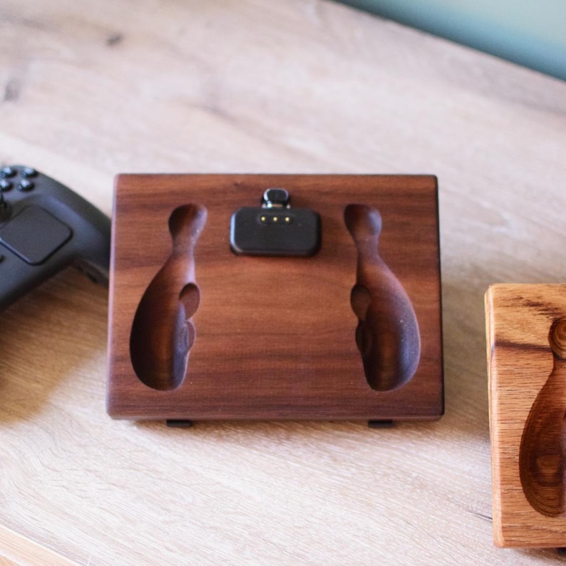 Pu&ograve; includere: Una base di ricarica rettangolare in legno marrone scuro con due rientranze intagliate per le impugnature del controller e un connettore di ricarica elettronico nero centrale. La base &egrave; posizionata accanto a un controller di gioco nero.