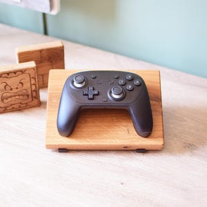Op de afbeelding: Een zwarte gamecontroller ligt op een lichtbruine houten standaard. De controller heeft meerdere knoppen en joysticks. Twee houten blokken met gebeeldhouwde gezichten staan op de achtergrond. De standaard is rechthoekig en eenvoudig.