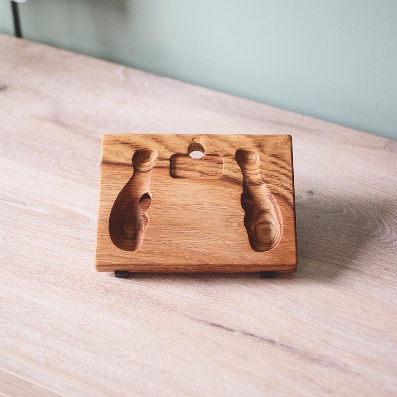 Pu&ograve; includere: Un supporto rettangolare in legno con venature naturali, due rientranze verticali intagliate e un foro circolare centrale sul bordo superiore. Il supporto poggia su due piccoli piedini neri alla base, offrendo una superficie di appoggio inclinata.