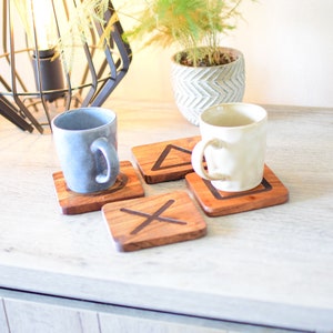 Op de afbeelding: Vier houten onderzetters met elk een ander controllersymbool. De symbolen zijn cirkel, driehoek, vierkant en X. Twee mokken staan op de onderzetters.
