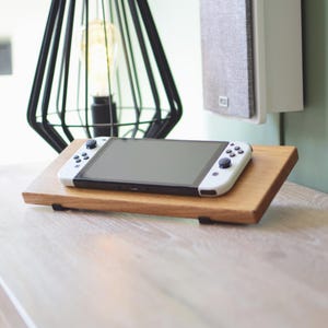 Op de afbeelding: Een witte en zwarte handheld gameconsole ligt op een kleine, schuine houten standaard. De standaard is lichtbruin en heeft zwarte steunen. Op de achtergrond staan een zwarte geometrische lamp en een witte luidspreker.