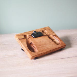 Pu&ograve; includere: Una base di ricarica rettangolare in legno con finitura a venature naturali. La superficie presenta due incavi intagliati a forma di birilli da bowling e un componente di ricarica elettronico nero centrale con tre piccoli pin di contatto dorati.