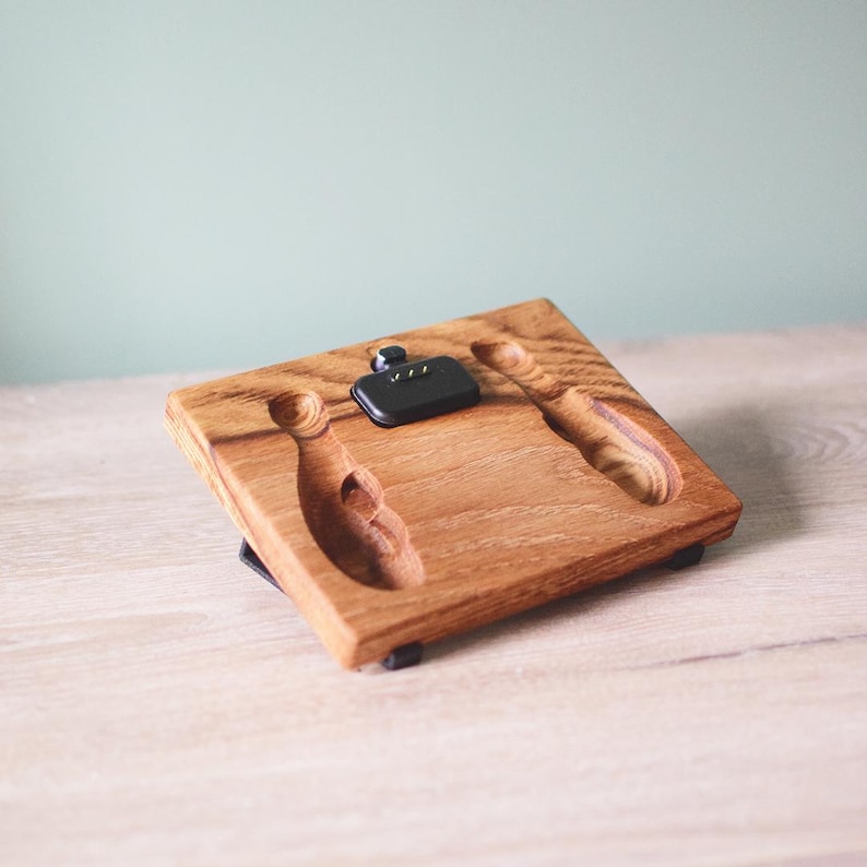 Pu&ograve; includere: Una base di ricarica rettangolare in legno con finitura a venature naturali. La superficie presenta due incavi intagliati a forma di birilli da bowling e un componente di ricarica elettronico nero centrale con tre piccoli pin di contatto dorati.