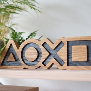 Op de afbeelding: Houten wanddecoratie met de iconische PlayStation-controller symbolen: driehoek, cirkel, kruis en vierkant. De symbolen zijn in hout gesneden en zwart geverfd.