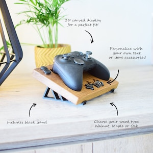 Op de afbeelding: Een houten standaard met een 3D-gesneden ontwerp voor een videogamecontroller. De standaard is gemaakt van walnoot, esdoorn of eikenhout. Het bevat een zwarte standaard en kan worden gepersonaliseerd met tekst of accessoires.