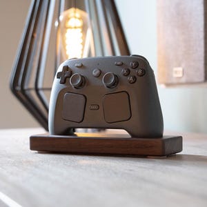 Può includere: Un controller per videogiochi grigio scuro con due levette analogiche, un pad direzionale e quattro pulsanti d'azione con lettere, poggiato su un supporto rettangolare in legno marrone scuro. Il controller presenta un logo centrale e due grandi touch pad.