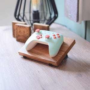 Op de afbeelding: Een mintgroene en witte gamecontroller met rode knoppen rust op een houten standaard. De controller heeft een afgerond ontwerp en is iets omhoog gekanteld. De standaard is gemaakt van lichtbruin hout.