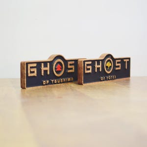 以下が含まれることがあります： 「GHOST」と「OF TSUHIMA」と「OF YOTEI」の文字が黒字で書かれた2つの木製看板。看板は茶色の縁取りで、赤と黄色のエンブレムが特徴です。看板は木製の表面にあります。
