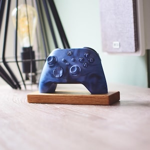 May include: A blue and white marbled Xbox controller sits on a small, rectangular wooden stand. The controller has various buttons and directional pads. The stand is made of light brown wood. A decorative lamp is in the background.