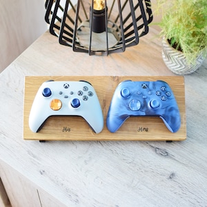 Może przedstawiać: Drewniany stojak z dwoma kontrolerami Xbox, jeden jest biały, a drugi niebieski z marmurowym wzorem. Stojak ma wygrawerowane słowa "His" i "Hers".