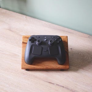 Pu&ograve; includere: Un controller per videogiochi nero con due levette analogiche, un pad direzionale e quattro pulsanti con lettere poggia su un supporto rettangolare in legno. Il controller presenta un logo centrale e due grandi touchpad sulla parte anteriore.
