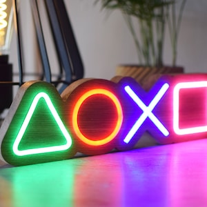 May include: A wooden sign with four neon lights shaped like the PlayStation controller symbols: a green triangle, a red circle, a blue X, and a pink square.