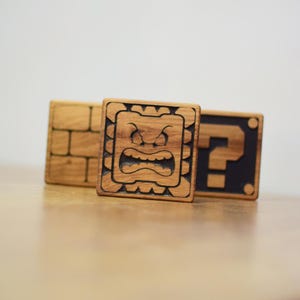 Peut inclure: Blocs en bois avec des motifs inspirés d'un jeu vidéo. L'un des blocs présente un motif de briques, un autre un visage en colère et le troisième un point d'interrogation. Les blocs sont en bois avec une finition naturelle.