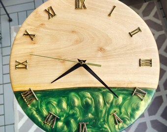 May include: A round wooden clock with Roman numeral markings and gold-colored hands. The bottom half of the clock face is made of green epoxy resin with gold flecks.