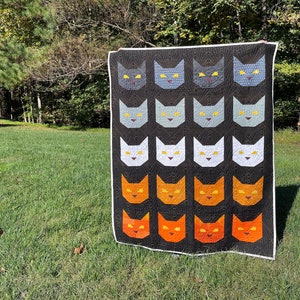 Könnte beinhalten: Eine schwarze Steppdecke mit einem sich wiederholenden Muster aus orangefarbenen, weißen und grauen Katzenköpfen mit gelben Augen. Die Steppdecke hängt an einer Wäscheleine in einem grasbewachsenen Bereich.
