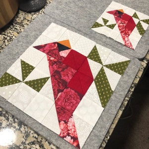 May include: Two quilt blocks featuring a cardinal design. The larger block has a red cardinal with floral patterned fabric, surrounded by white and green geometric shapes. The smaller block mirrors the design. The blocks are on a gray surface.
