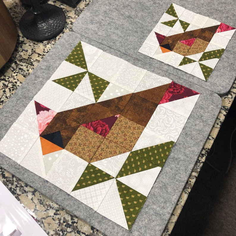 May include: Two bird-themed quilt blocks in progress. The larger block features a brown bird with floral and geometric patterns. The smaller block mirrors the design. Both are on gray ironing mats, showcasing the patchwork details and color palette.
