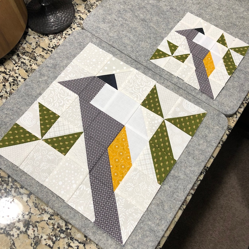 May include: Two quilt blocks featuring a bird design. The birds have gray, white, and yellow accents. The blocks are on a gray surface. The bird's body is made of various patterned fabrics.