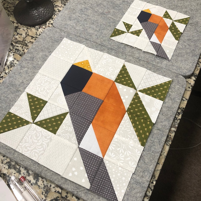 May include: Two bird-themed quilt blocks. The larger block features a bird design with orange, black, white, and green geometric shapes. The smaller block mirrors the design. Both are on a gray surface, showcasing a patchwork style.