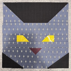 May include: A black and blue patchwork quilt block with a cat design. The cat has yellow eyes and a burgundy nose.