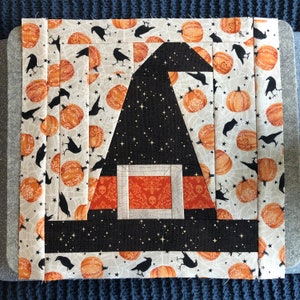 May include: A black and white witch hat quilt block with a white background featuring a repeating pattern of orange pumpkins, black crows, and spiderwebs. The hat has a black brim and a black crown with white stars.
