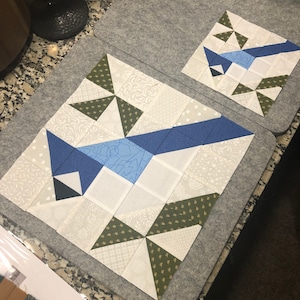 May include: Two fabric quilt blocks featuring a blue jay design. The larger block has a blue jay with blue, white, and green triangular accents. The smaller block mirrors the design. The blocks are on a gray surface.