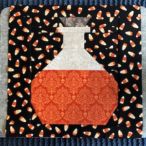 May include: A patchwork fabric design of a potion bottle with a black background and candy corn pattern. The bottle is filled with a red liquid and has a white web-like design.