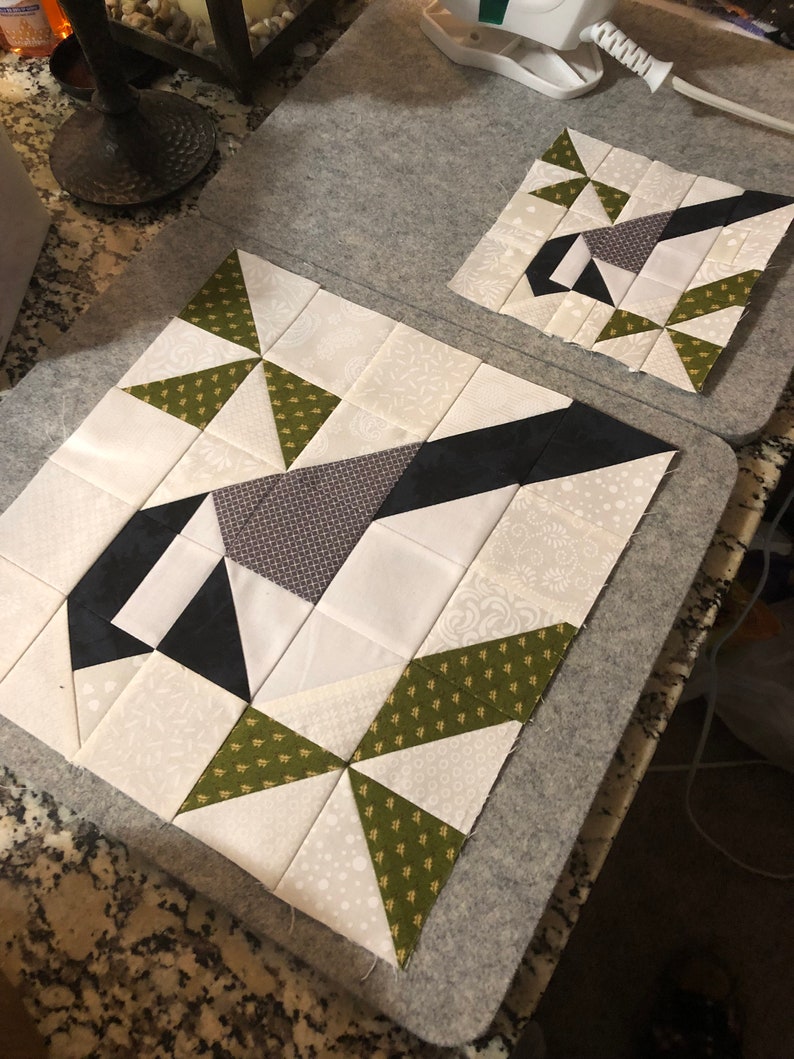 May include: Two quilt blocks in progress, featuring geometric patterns in white, black, gray, and olive green fabrics. The blocks are on a gray ironing board, showcasing the patchwork design and quilting process.