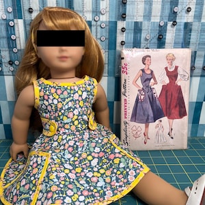 Puede incluir: Una muñeca con un vestido estampado floral con ribetes y botones amarillos. El vestido tiene un fondo azul oscuro con flores rosas, amarillas y blancas. La muñeca lleva zapatos blancos. Un patrón de costura vintage está en el fondo.
