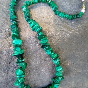 May include: A green malachite gemstone necklace with irregular shaped beads. The necklace is on a gray background.