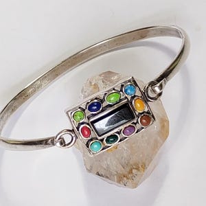 May include: A silver-toned metal bangle bracelet featuring a rectangular centerpiece. The center holds a black rectangular stone surrounded by twelve small, oval-shaped cabochon stones in various colors including red, green, blue, and yellow.