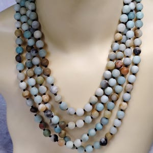 May include: A multi-strand necklace featuring numerous round, matte-finish beads in shades of pale blue, soft green, cream, and earthy brown. The beads are strung on a thin black cord, creating a layered effect that drapes across the neck area.