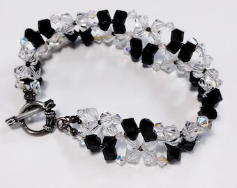 May include: A bracelet constructed from alternating black and clear faceted crystal beads arranged in a repeating pattern. The piece features a circular silver-toned toggle clasp with a textured finish, connecting the two ends of the beaded band.