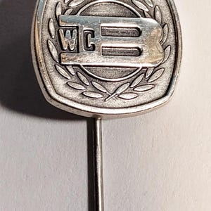 May include: A silver-toned lapel pin with a square, rounded edge design. The pin features the letters "WC" above a stylized "B" within a laurel wreath. The pin has a long, thin, silver-colored stem.