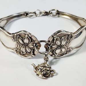 May include: A silver-toned bracelet crafted from two curved spoon handles featuring embossed floral patterns. The two ends are connected by a circular jump ring that holds a small, three-dimensional silver-toned teapot charm with floral details.