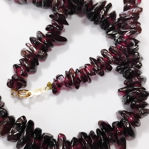 May include: A necklace composed of dark red, irregularly shaped garnet stones. The stones are strung together, forming a continuous loop. A gold-colored clasp secures the necklace. The gemstones have a polished, glossy appearance.